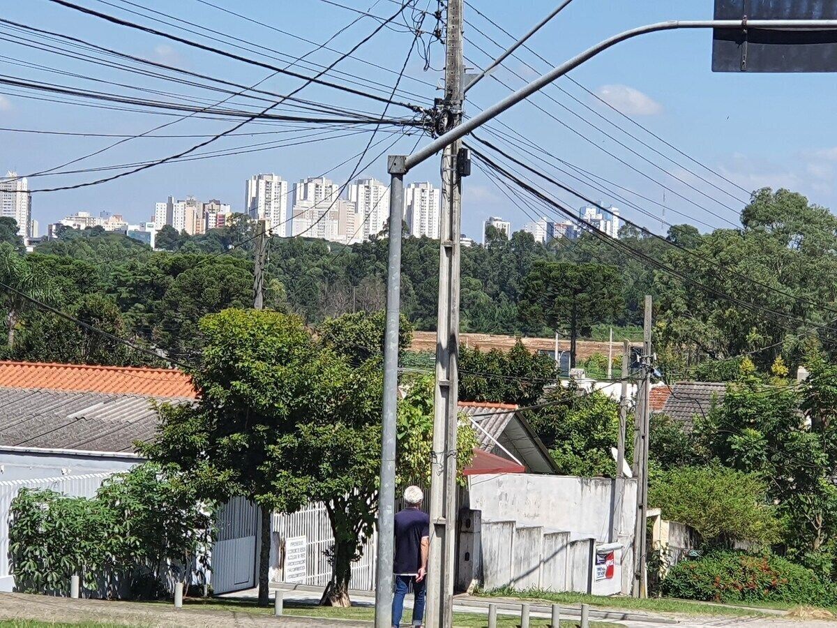 A cityscape with buildings, a person standing near a utility pole, and a fenced area with greenery.