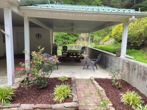 Une terrasse couverte, dotée d’une table et de chaises, et entourée d’un jardin agrémenté de plantes et de paillis.