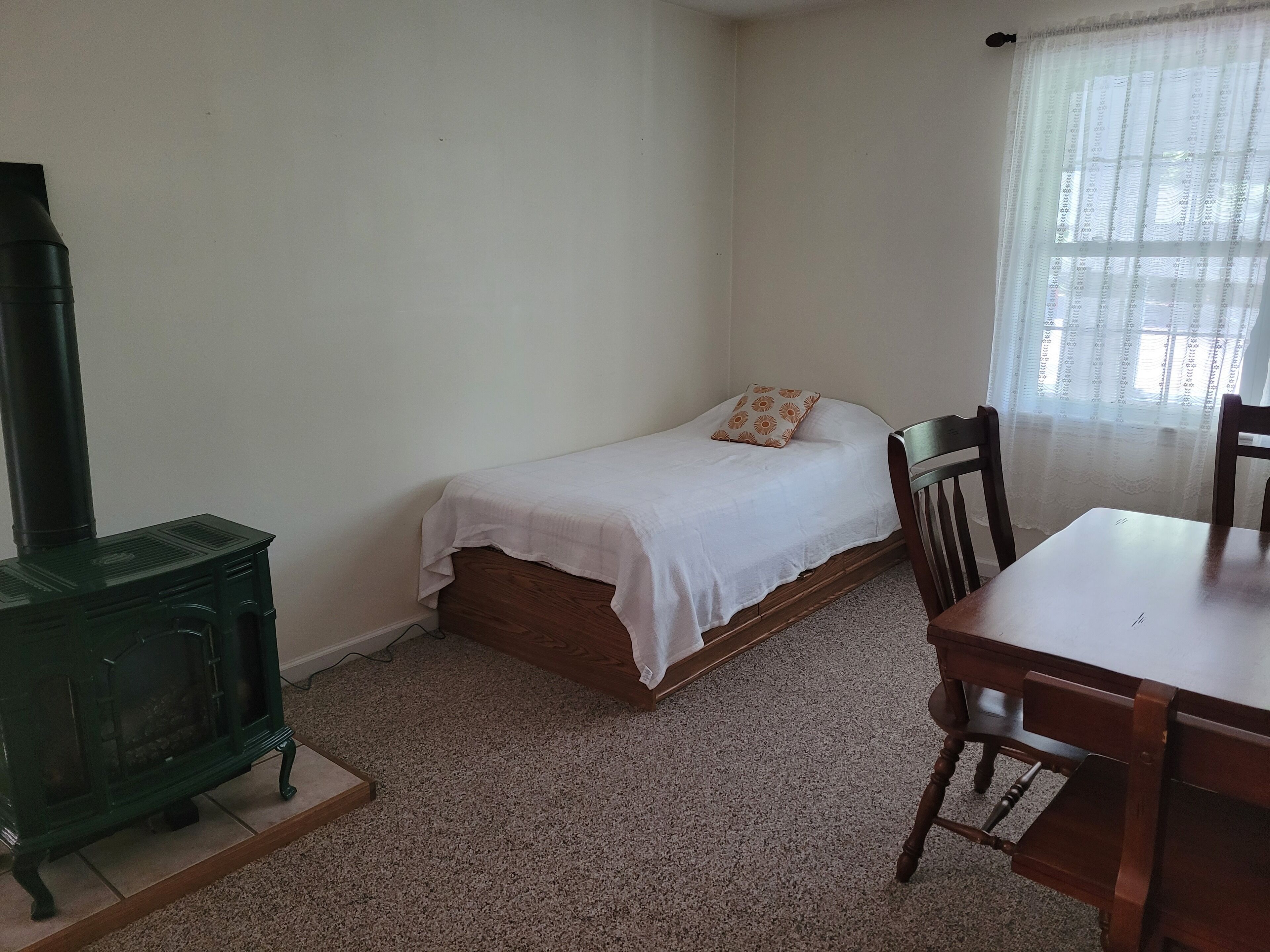 Una habitación pequeña y poco amoblada, con una cama, una mesa de madera, una silla y una estufa verde.