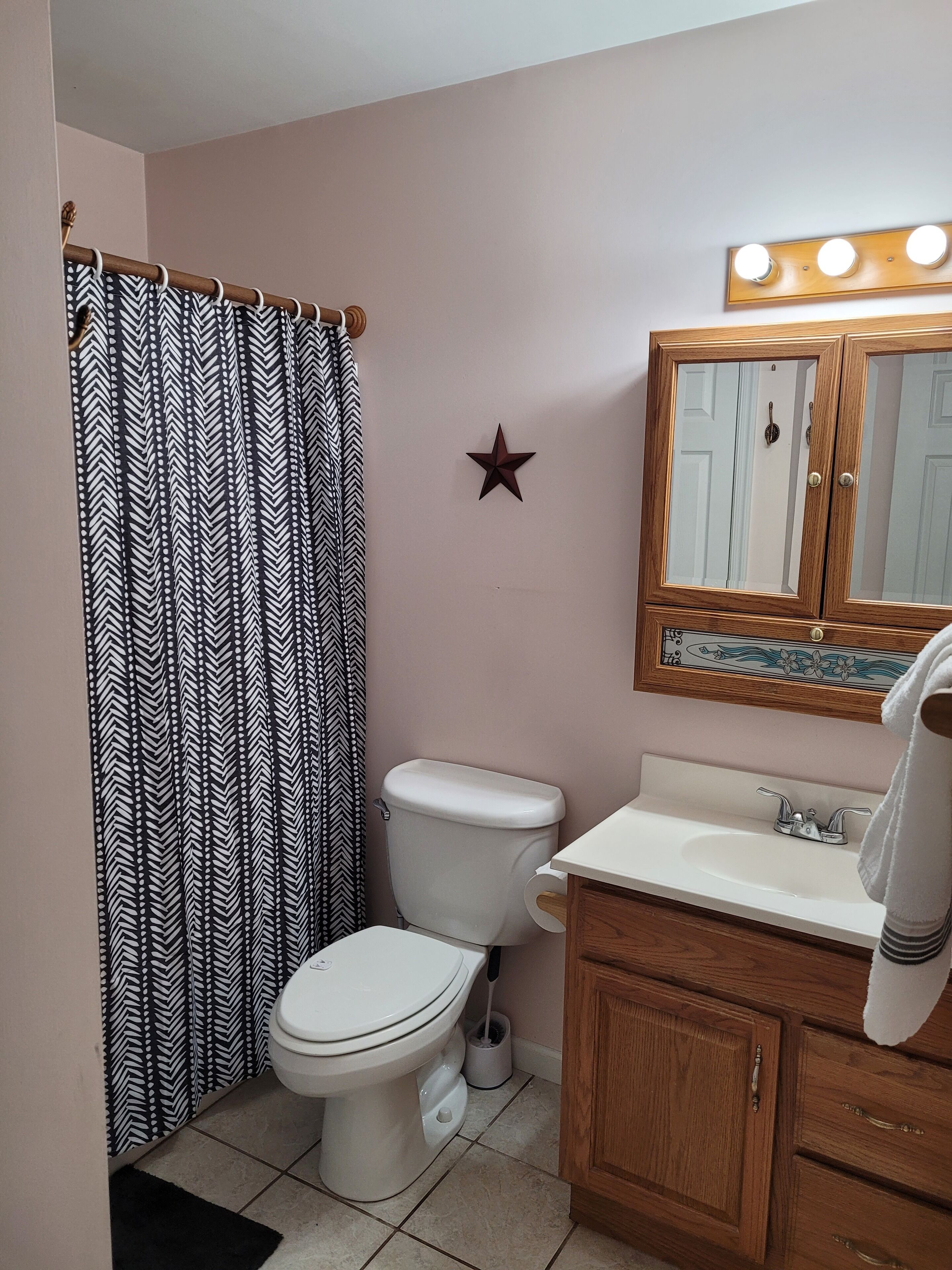 A bathroom with a white toilet, a shower curtain with a black and white chevron pattern, a star decoration on the wall, a wooden cabinet with a mirror above the sink, and a towel hanging on the cabinet.