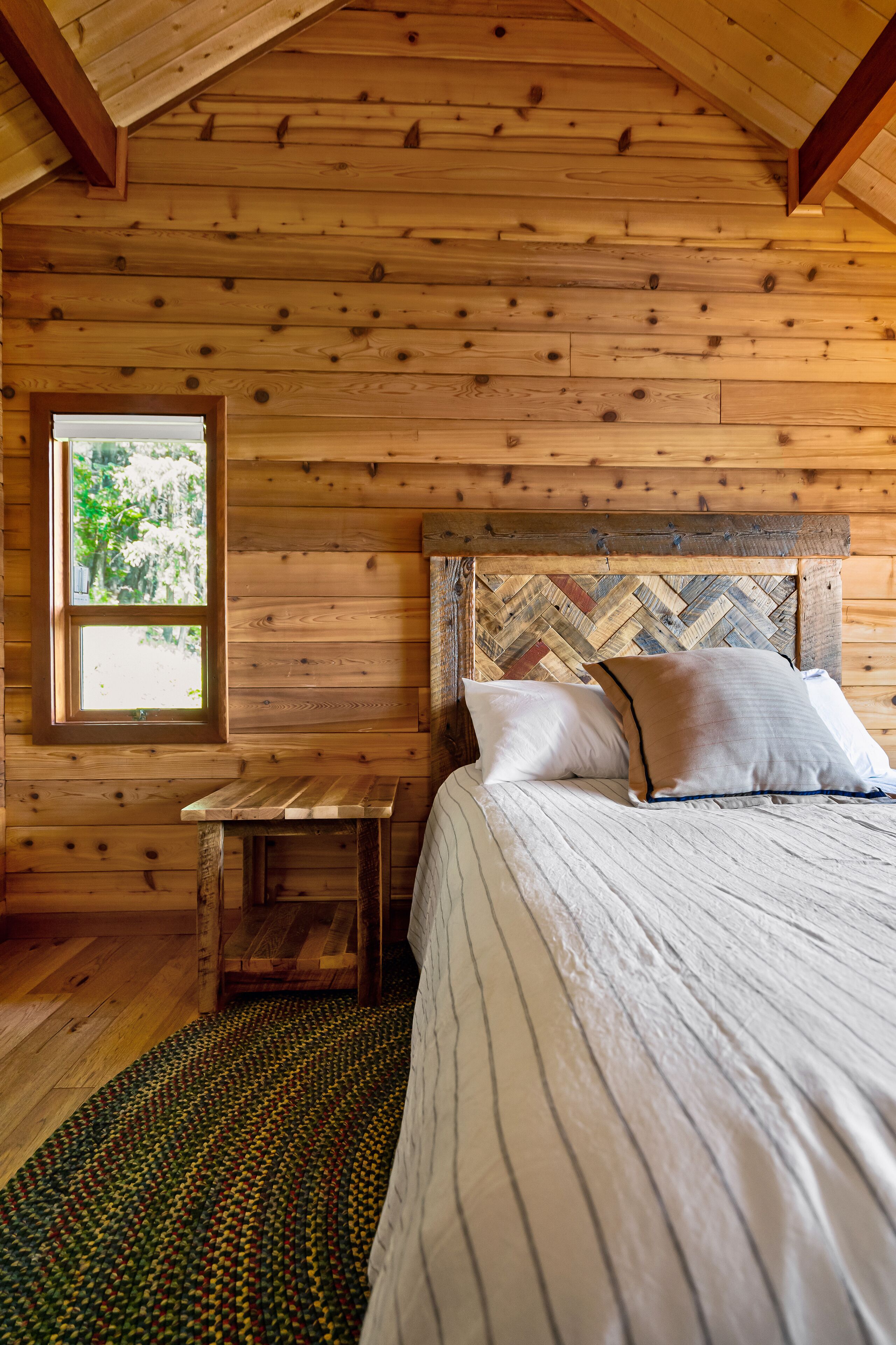 Un dormitorio de cabaña de madera con cama, almohadas, una ventana y una pequeña mesa.