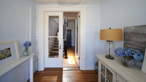 Un pasillo con suelo de madera, una puerta blanca que da a una escalera y una lámpara sobre una mesita consola.