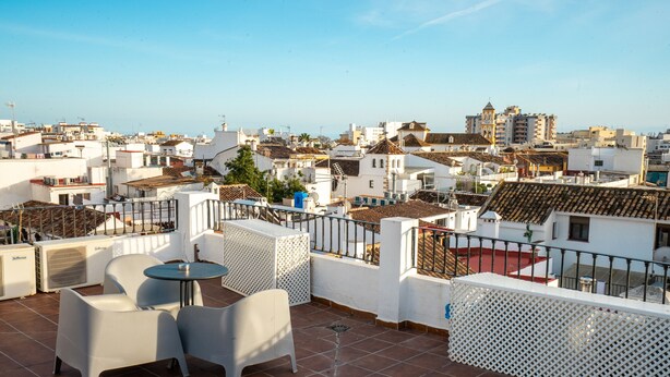 A rooftop terrace with white chairs and a small table, overlooking a cityscape with white buildings and a clear sky. - Marbella Village  | Studio with Juliette Balcony (Marbella)