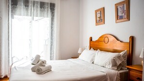 A neatly made bed with white linens and rolled towels, a wooden headboard, a bedside table with a lamp, and two framed pictures on the wall. - Marbella Village  | Studio with Juliette Balcony (Marbella)