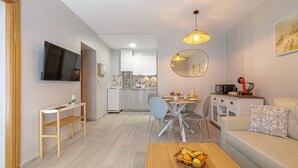 A modern living room with a sofa, a coffee table with fruit, a dining area with chairs, a kitchen in the background, and a TV mounted on the wall.