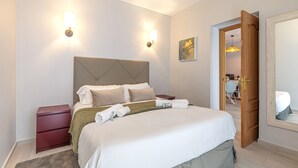 A neatly made bed with pillows, a nightstand, a wooden door, and a wall-mounted light fixture.