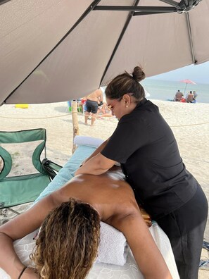 Montagem para massagem à beira-mar, com mesa de massagem, uma cadeira verde e uma tenda para dar sombra.