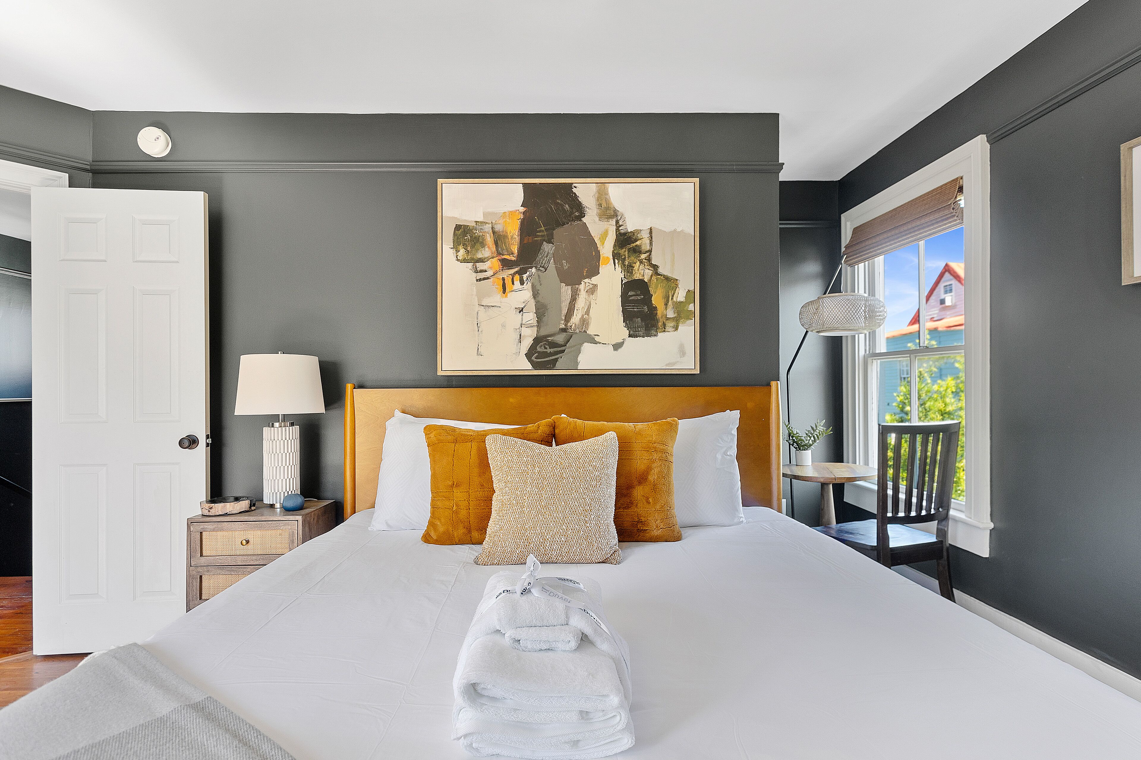 A neatly made bed with a white comforter and two pillows, a bedside table with a lamp, and a painting above the bed.