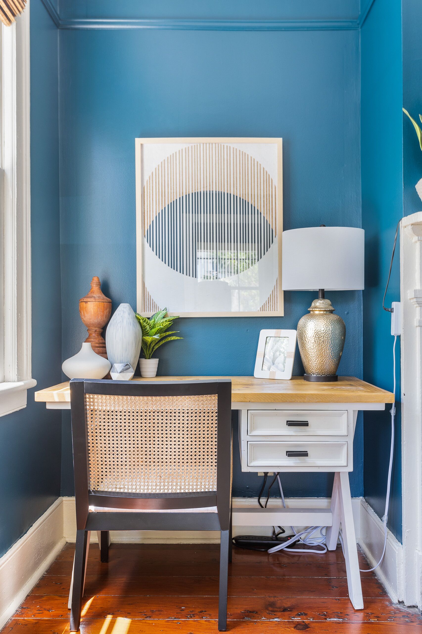 A room with a blue wall, a wooden desk, a wicker chair, a white drawer unit, a framed artwork, a vase, a potted plant, and a lamp.
