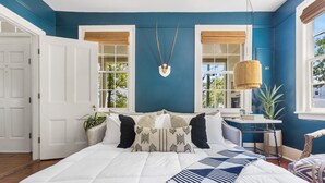 A bedroom with a large bed, a headboard with a decorative item, a bedside table with a lamp, and a view of the outdoors through a window. - Steps Away from Historic King! | The Sutton - B (Charleston)