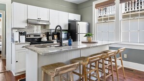 A kitchen with white cabinets, a stainless steel refrigerator, a granite countertop, and wooden bar stools. - Steps Away from Historic King! | The Sutton - B (Charleston)