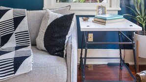 A room with a blue wall, a white window with a checkered blind, a hanging lamp, a sofa with patterned cushions, a small table with books, and a potted plant. - Steps Away from Historic King! | The Sutton - B (Charleston)