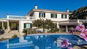 A large, rectangular swimming pool with a clear blue water surface, surrounded by a white fence. In the background, there is a two-story building with a balcony and a tree in front. - Architectural jewel between sea and mountains (Teià)