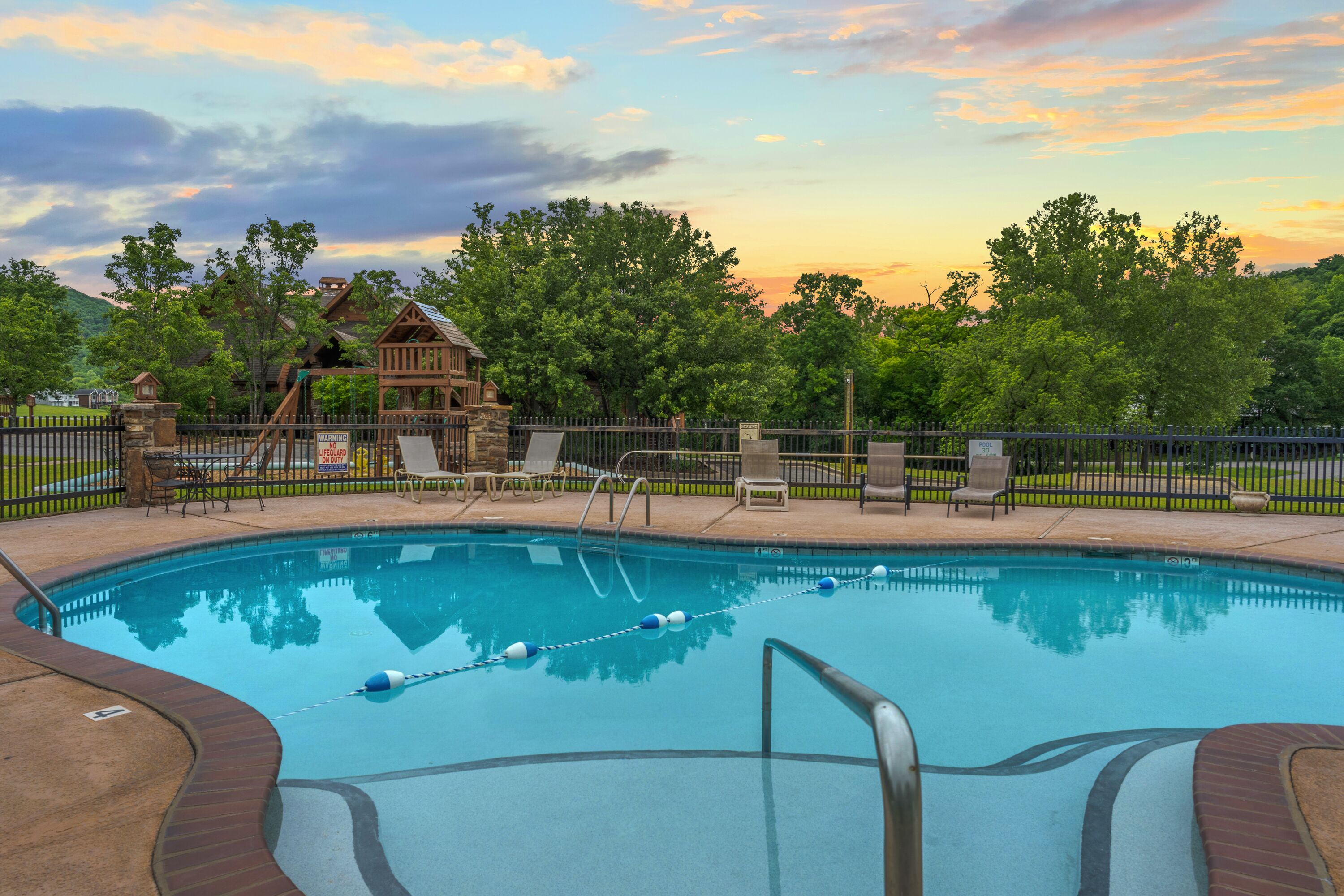 A swimming pool with a wooden deck, lounge chairs, and a fenced area with a view of trees and a building in the background.