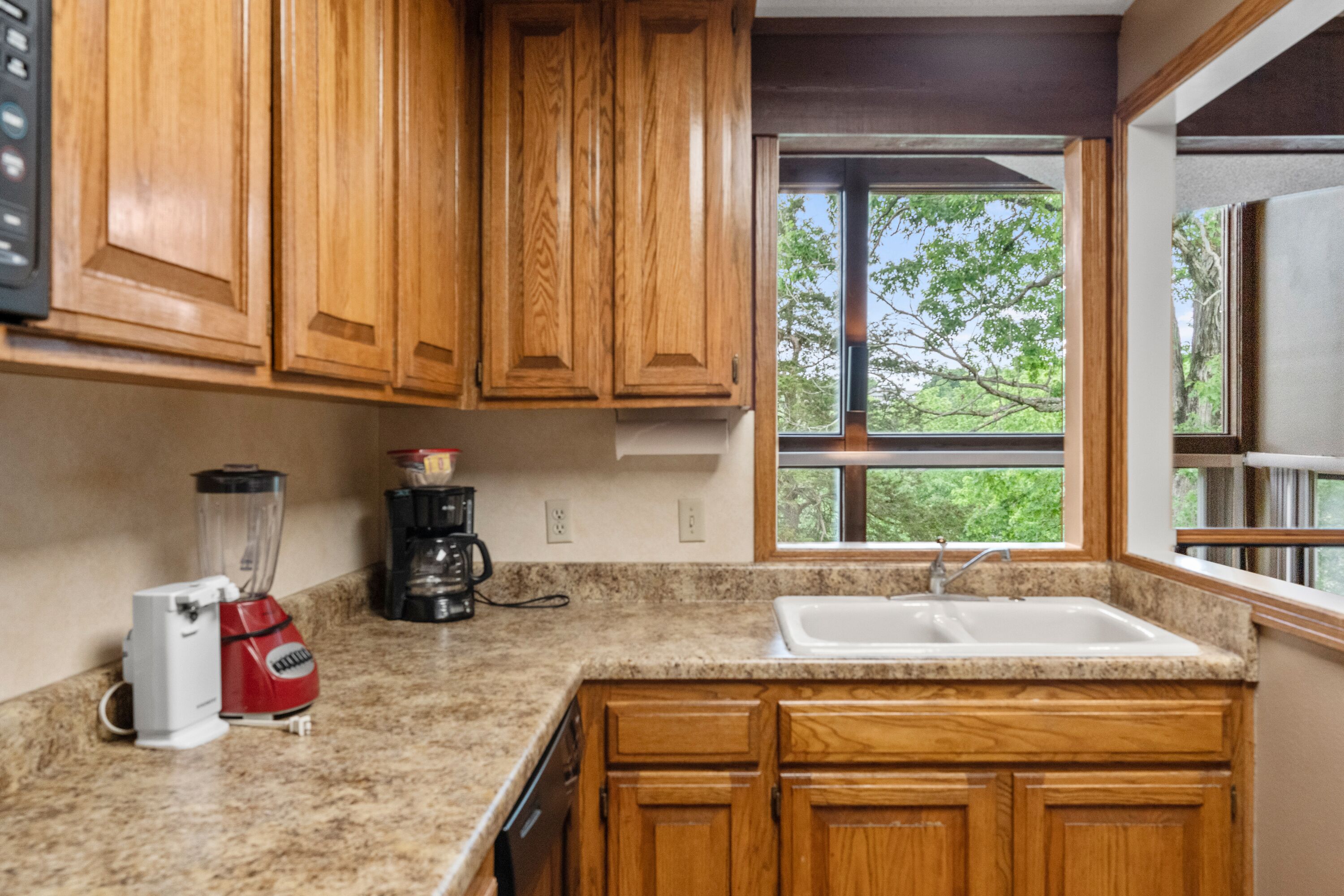 Eine Küche mit Holzausstattung, einer Arbeitsplatte aus Granit, einem Mixer, einer Kaffeemaschine und einem Fenster mit Blick auf Bäume.