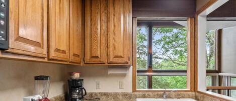 A kitchen with wooden cabinets, a granite countertop, a blender, a coffee maker, and a window with a view of trees.
