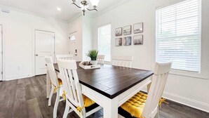Een eetkamer met een donker houten tafel, witte stoelen met gele kussens, een kroonluchter en ingelijste foto's aan de muur.