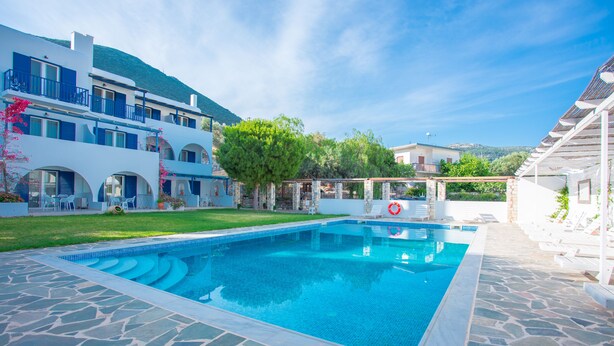 Una piscina con baldosas de piedra, una zona verde y un edificio con varios balcones y una pérgola.