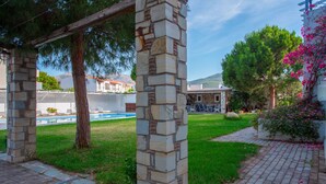 Una estructura de pérgola de piedra en un patio con piscina, follaje y un edificio con techo rojo al fondo.