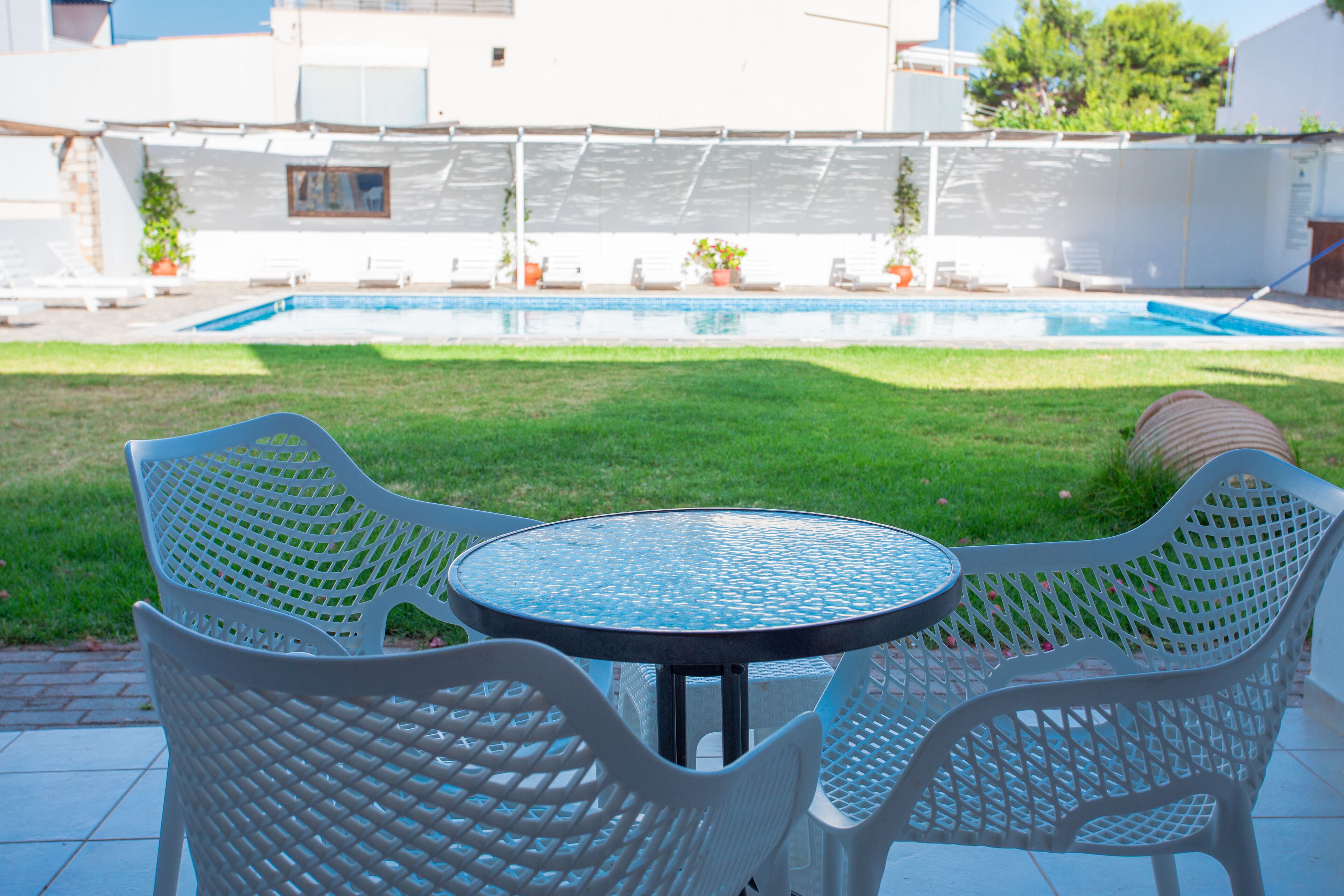 A patio with a round table and two white chairs, a swimming pool, and a white building in the background.