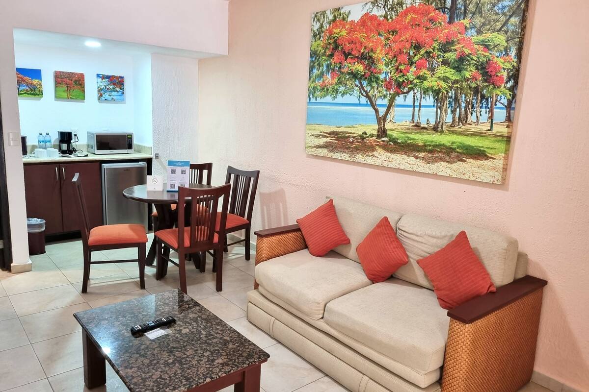 A living room with a sofa, chairs, a coffee table, a dining area with a table and chairs, a refrigerator, and a painting of a tree with red flowers.