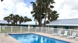 A swimming pool with white lounge chairs and a white fence, surrounded by palm trees and a building with a blue roof. - Captain Quarters Motel (Port O Connor)