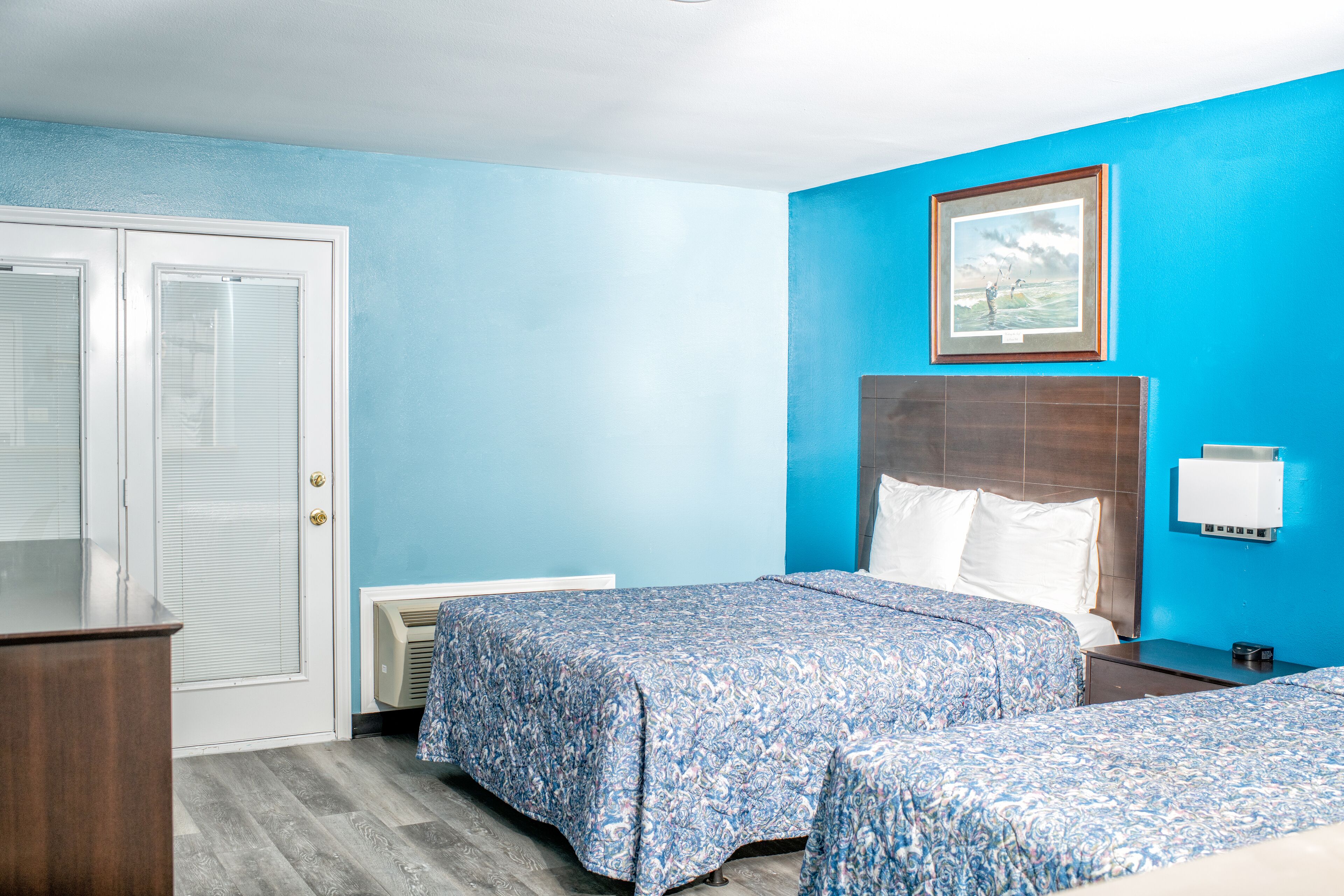 Une chambre d’hôtel avec deux lits, une porte blanche, un tableau encadré au mur et une table de chevet avec une lampe.