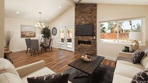 Een ruime woonkamer met een open haard, een eethoek en een groot raam met uitzicht op palmbomen.