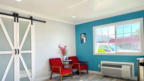 A room with a sliding glass door, two red chairs, a small table with a vase of flowers, a window with a view of cars, and a radiator. - Motel 6 Yuba City, CA (Yuba City)