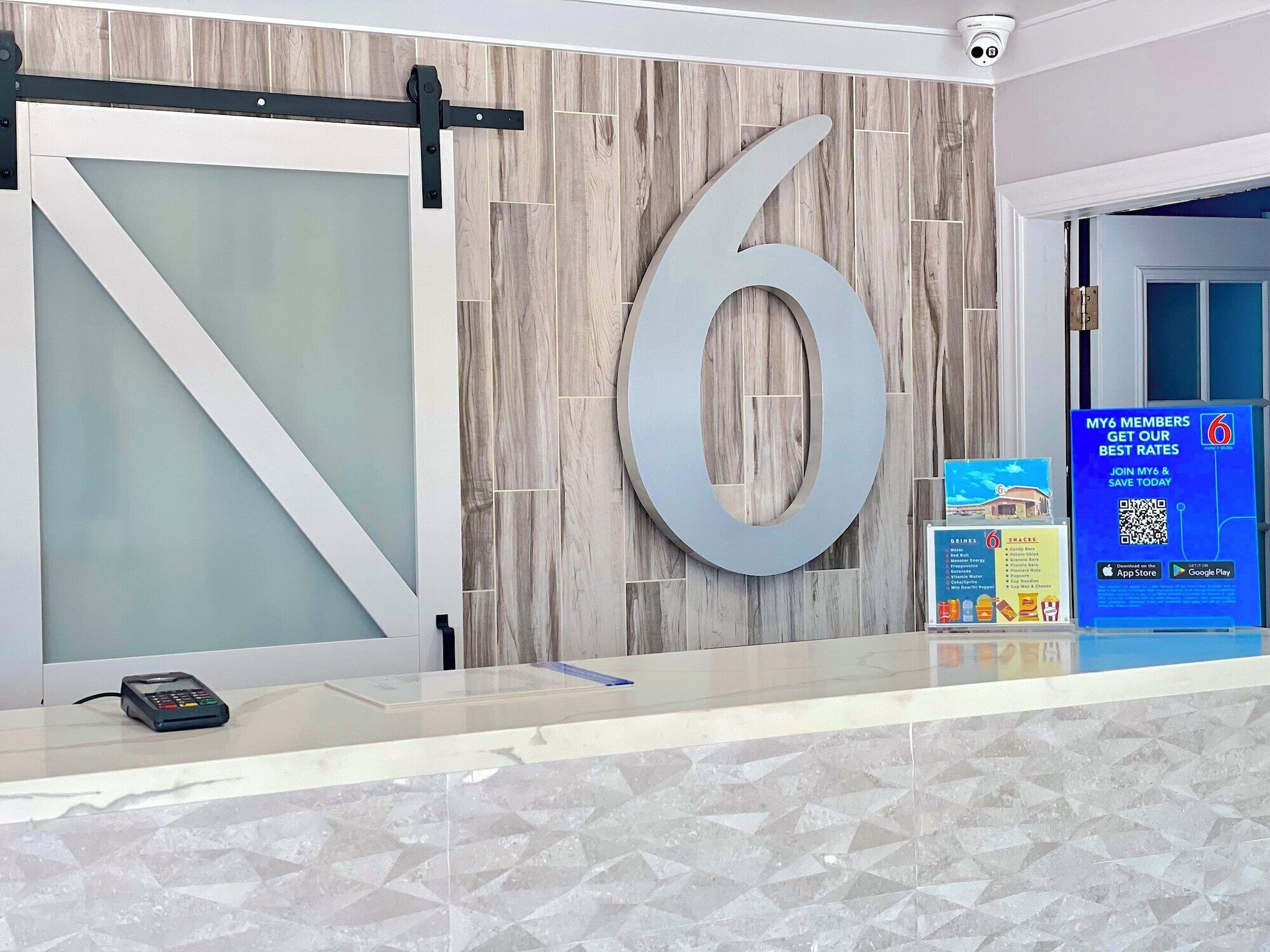 A reception desk with a large number '6' on the wall, a security camera, and a QR code sign advertising My6 membership rates.