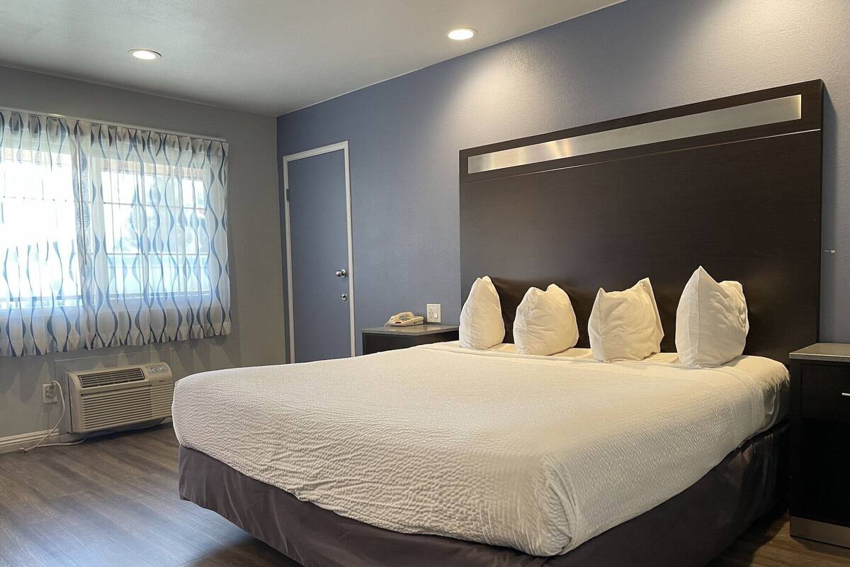 A modern hotel room with a large bed, a bedside table, a nightstand, a wall-mounted light, and a window with curtains.