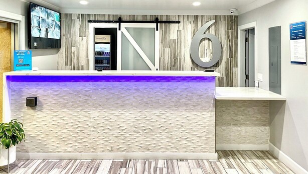 A reception area with a modern design, a large number '6' sign, a television mounted on the wall, and a reception desk with a blue light. - Motel 6 Yuba City, CA (Yuba City)