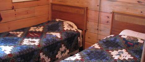 A cabin-style room with two single beds, wooden walls, a wooden dresser, and a wall-mounted lamp.