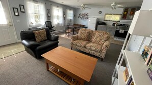 Una sala de estar con un sofá de cuero negro, un sofá estampado con motivos florales y una mesa de centro de madera. Hay un comedor con mesa y sillas, y una cocina con electrodomésticos y armarios.