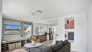 Una sala de estar moderna con sofá, mesa de centro y televisor. Se observa una zona de cocina con armarios y refrigerador.