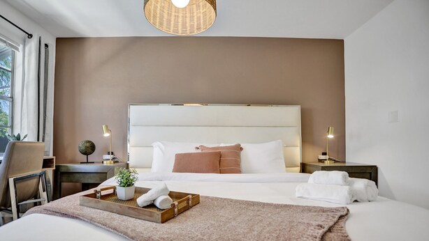 Habitación de hotel moderna con una cama grande, una mesita de noche con lámpara, una silla y una ventana con cortinas.