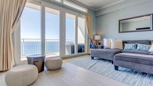A spacious room with a large sliding glass door leading to a balcony, a grey sofa set, and a round ottoman. - The Lazy Place: Gulf Penthouse, High Ceiling &Bunk (Panama City Beach)