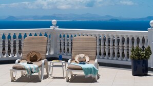 Dos sillones de descanso con sombreros y una manta, una pequeña mesa con una botella y una planta en maceta en un balcón con barandilla blanca y vistas al mar y a las montañas.