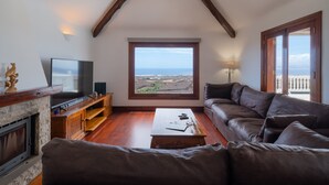 Una sala de estar con chimenea de piedra, televisor, mueble de entretenimiento de madera, sofá de cuero marrón, mesa de centro y un ventanal con vistas al paisaje.