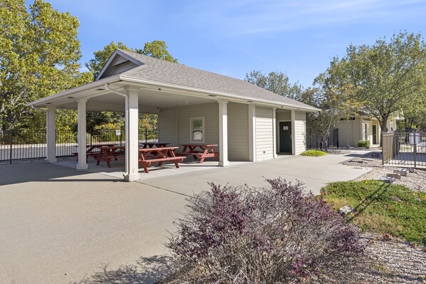 A covered picnic area with benches and a small building, surrounded by trees and a fence.