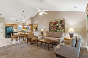 Ein Wohnzimmer mit einer Couch, einem Couchtisch und Sesseln, ein Essbereich mit Tisch und Stühlen, sowie eine Küche mit Schränken und Geräten.