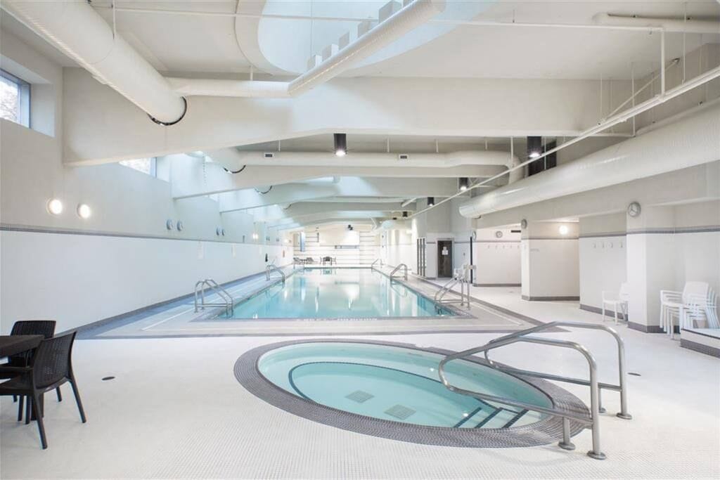 Ein großer, überdachter Schwimmbereich mit Sprungtürmen in der Mitte, umgeben von Sitzgelegenheiten und einer hohen Decke mit freiliegenden Leitungen.