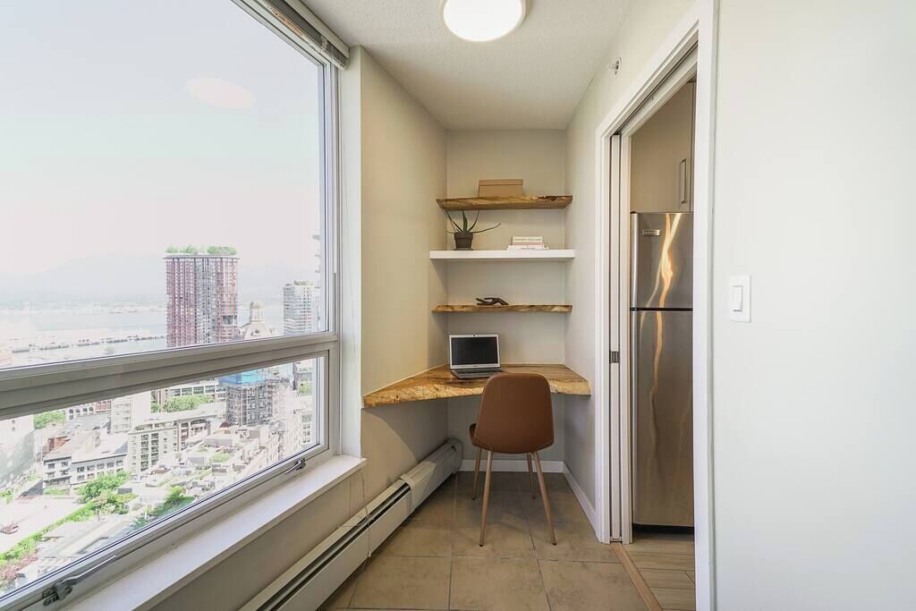 A small kitchen with a built-in refrigerator, a laptop on a wooden shelf, and a chair.