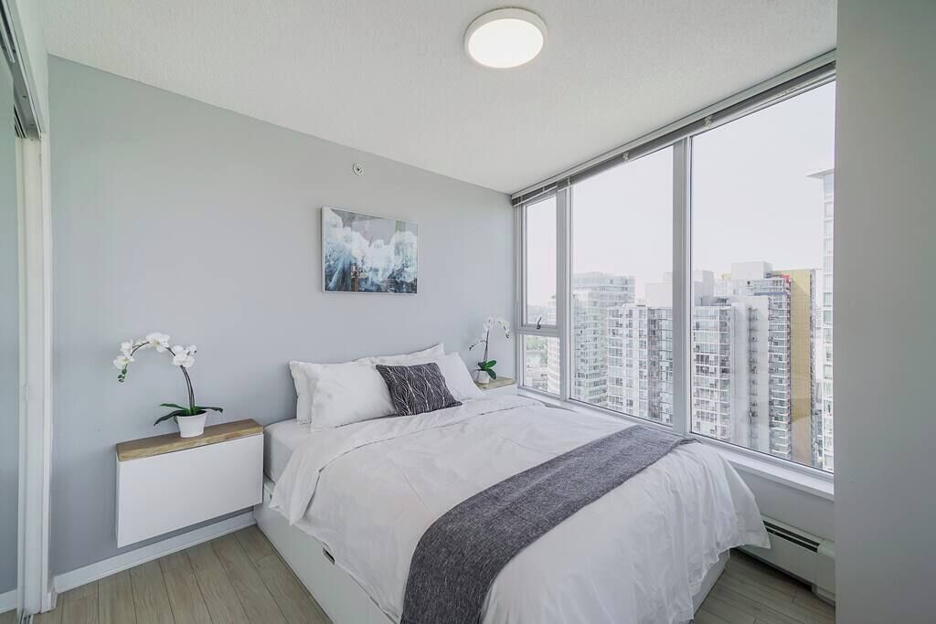 Ein modernes Schlafzimmer mit großem Fenster, einem Bett mit weißer Bettwäsche, einem Nachttisch mit Pflanze und Blick auf die Stadt.