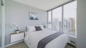 A modern bedroom with a large window, a bed with white bedding, a nightstand with a plant, and a city view.