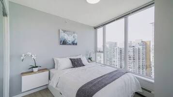 Ein modernes Schlafzimmer mit großem Fenster, einem Bett mit weißer Bettwäsche, einem Nachttisch mit Pflanze und Blick auf die Stadt.