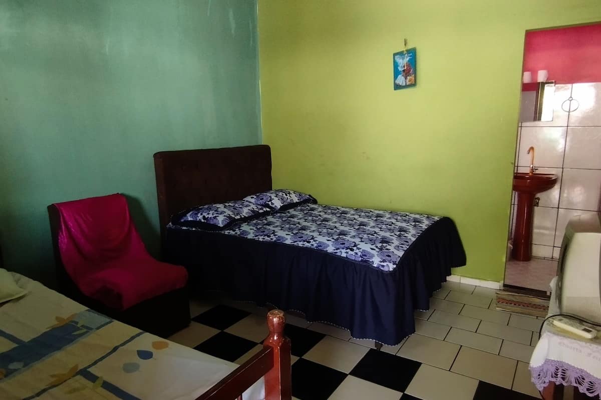 A small, single-room accommodation with a bed, a red chair, a bedside table with a lamp, and a small sink.