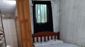 Una habitación pequeña con baldosas, que incluye una cama, una silla de madera, un inodoro y una ventana con cortinas.