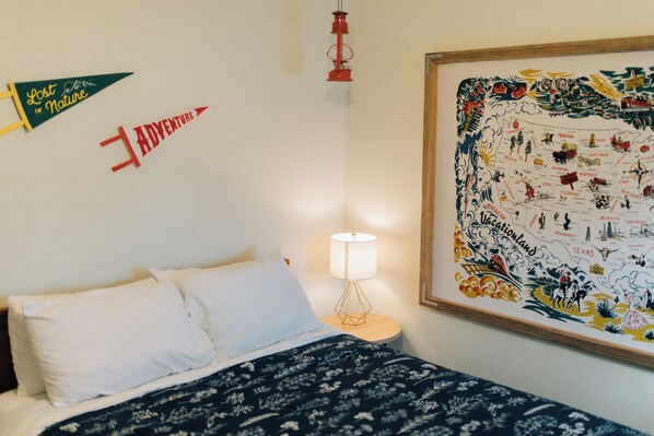Un lit bien fait, avec des oreillers blancs et une couverture à motifs sombres, une lampe de chevet et une grande carte murale des États-Unis indiquant les noms des États et les principaux points d’intérêt.