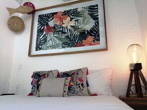 Un lit avec des coussins colorés, une œuvre d’art tropical encadrée, une prise de courant au mur et une lampe sur pied.